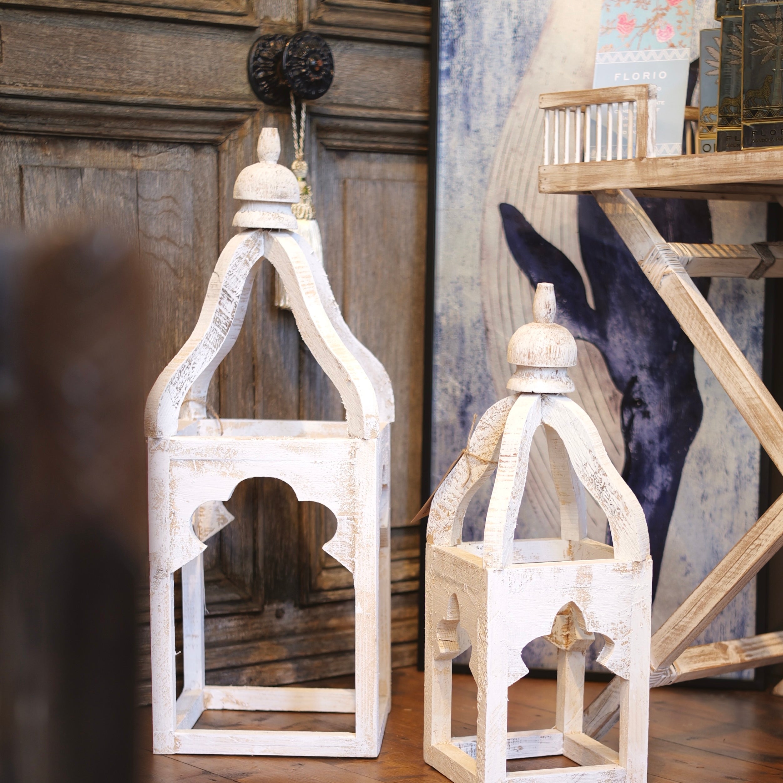 Two white wooden lanterns on a wooden floor with a decorative wall in the background.