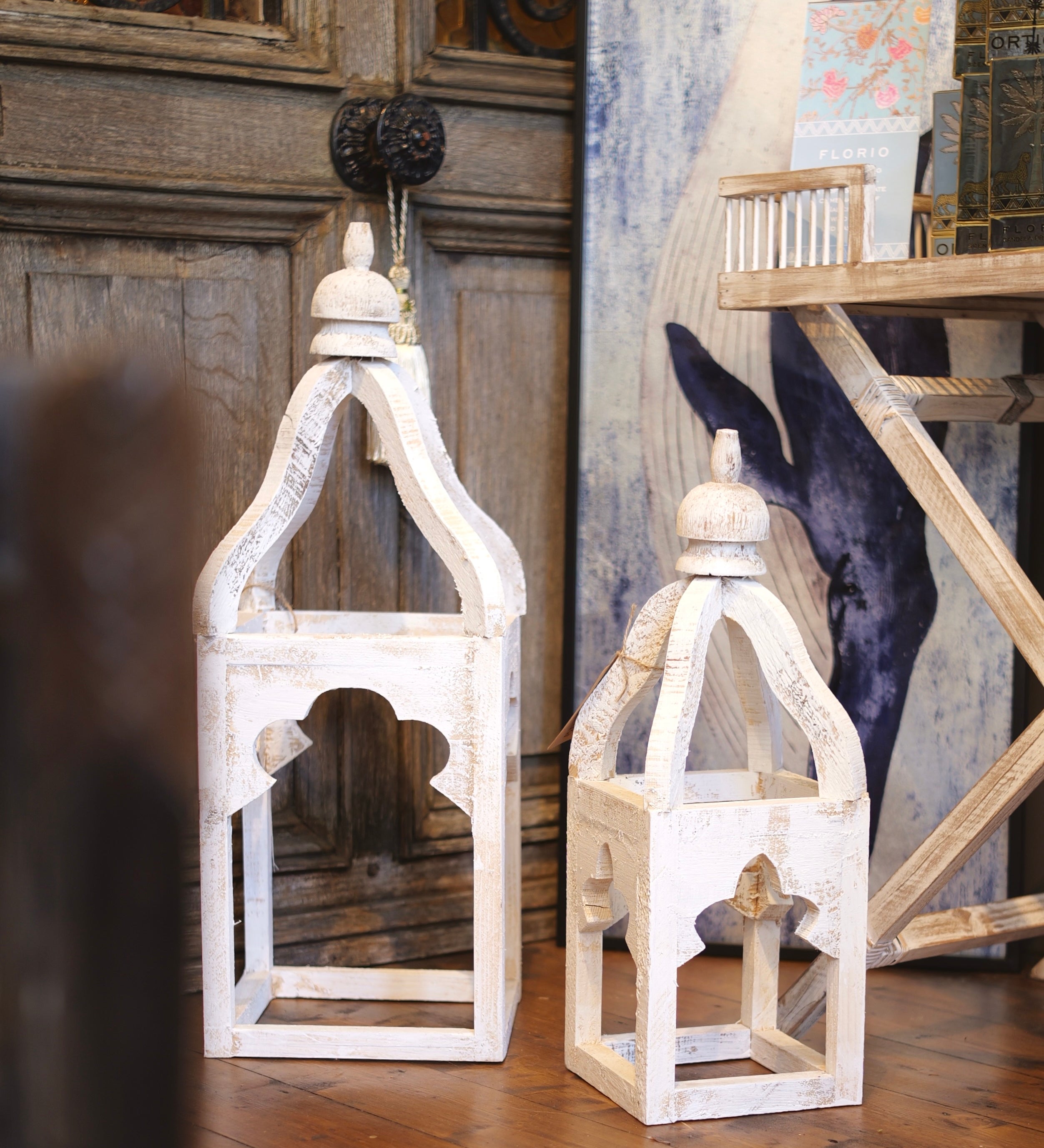 Two white wooden lanterns on a wooden floor with a decorative wall in the background.